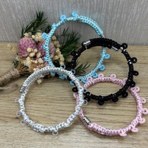 Puede incluir: Cuatro pulseras hechas a mano en varios colores: azul claro, negro, plateado y rosa. Cada pulsera presenta un diseño tejido con pequeños detalles decorativos. Las pulseras se exhiben sobre una superficie de madera.