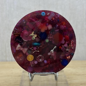 Könnte beinhalten: Ein kreisförmiges Kunstwerk aus Harz in tiefen Magenta- und Rosatönen, eingebettet mit verschiedenen Anhängern, darunter ein kleiner silberner Schlüssel, eine Goldmünze, eine hellrosa Schleife sowie diverse bunte Perlen, Glitzer und kleine Blumen.
