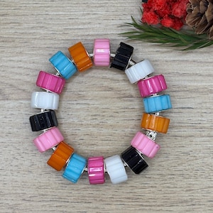 Pode incluir: Uma pulseira colorida com contas em tons de rosa, laranja, azul, preto e branco. As contas têm uma textura canelada e são enfiadas com metal prateado. A pulseira é exibida em uma superfície de madeira clara.