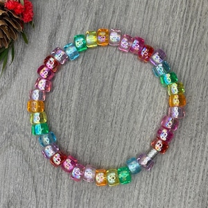 May include: A colorful beaded bracelet with a rainbow of hues, including pink, orange, yellow, green, and blue. The beads are square-shaped and strung together on a silver-colored elastic band. The bracelet is displayed on a gray wooden surface.