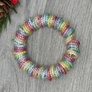 May include: A rainbow-coloured beaded hair tie in a circular shape. The hair tie is made of small, iridescent beads in a variety of colours. The beads are strung together to form a spiral. The hair tie is approximately 10 cm in diameter.