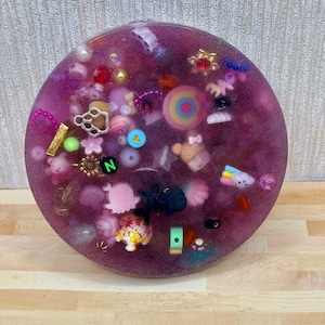 A circle shaped picky pad. The colour is purple. It is made from a reusable material and includes beads, charms, flatbacks and other embellishments.