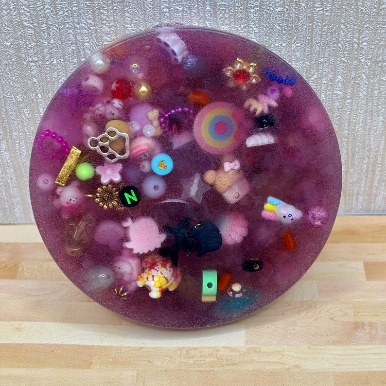 A circle shaped picky pad. The colour is purple. It is made from a reusable material and includes beads, charms, flatbacks and other embellishments.