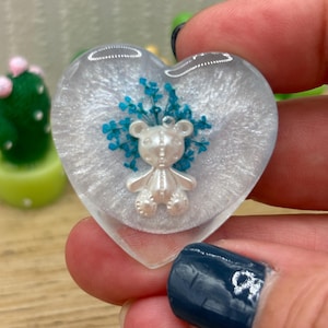 May include: Heart-shaped resin pendant with a pearlescent finish, featuring a small white teddy bear and blue floral accents. The pendant is clear around the edges, creating a layered effect. A unique, handcrafted accessory.