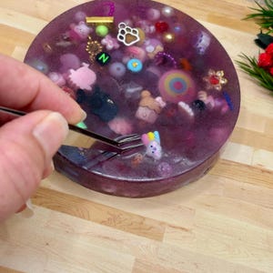 A circle shaped picky pad. The colour is purple. It is made from a reusable material and includes beads, charms, flatbacks and other embellishments.  Tweezers are grasping a small, colourful bunny charm.