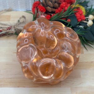May include: A clear, amber-coloured, decorative glass object with a bubbly, irregular surface. The object is round and sits on a light-coloured wooden surface. The background includes red and green floral elements.