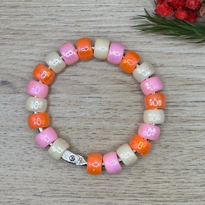 Pode incluir: Uma pulseira de contas com contas coloridas em tons de rosa, laranja e branco sujo. A pulseira é enfiada em um fio prateado e tem um fecho prateado. As contas têm aproximadamente 1,3 cm de comprimento.