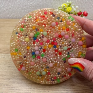 May include: A circular coaster made of clear resin embedded with a dense layer of small, cream-coloured spherical beads. The surface is accented with scattered, larger matte-finish spheres in vibrant shades of pink, yellow, green, blue, and red.
