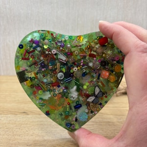 A green coloured picky pad in a heart shape. The sensory toy contains a variety of beads, charms, and other embellishments.
