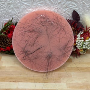 May include: A circular, pink item with black strands spread across it. It rests on a wooden surface, with floral decorations in the background. The item's purpose is unclear.