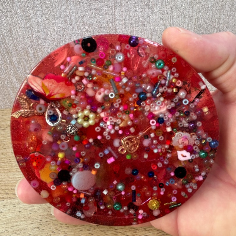 A circle shaped picky pad. The colour is pink. It is made from a reusable material and includes beads, charms, flatbacks and other embellishments.