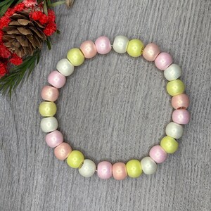 May include: A pastel-colored beaded bracelet featuring round beads in shades of pink, yellow, and white. The bracelet is displayed on a gray surface, with a decorative floral element in the upper left corner.