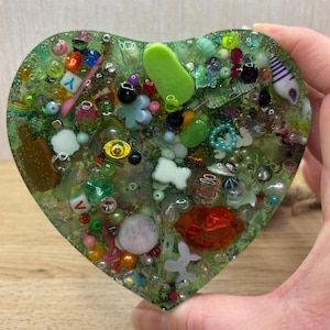 A green coloured picky pad in a heart shape. The sensory toy contains a variety of beads, charms, and other embellishments.