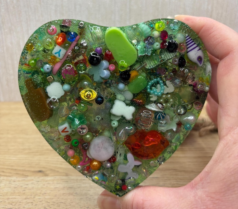 A green coloured picky pad in a heart shape. The sensory toy contains a variety of beads, charms, and other embellishments.