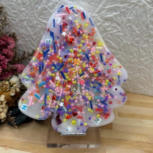 May include: A clear, tree-shaped resin ornament filled with colourful sprinkles, beads, and star-shaped confetti. The ornament has a white base and is displayed on a clear stand. The design includes red, blue, yellow, and pink elements.