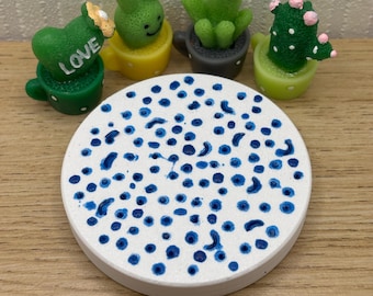 May include: A white, circular ceramic coaster decorated with blue dot patterns. Four small, decorative figurines of cacti and a bunny are arranged around the coaster. The figurines are green, yellow, and grey, with small flowers and the word "LOVE" on one.