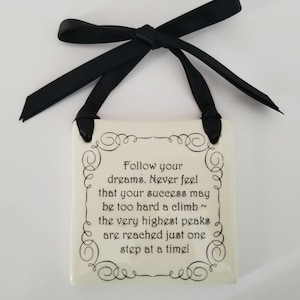 May include: A square, off-white ceramic plaque with black text and decorative swirls. The plaque is suspended by a black ribbon tied in a bow. The text reads: "Follow your dreams. Never feel that your success may be too hard a climb ~ the very highest peaks are reached just one step at a time!"