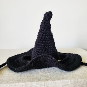 Pode incluir: Um chapéu de bruxa preto de crochê com uma coroa pontuda e uma aba larga. O chapéu é feito de um fio texturizado e tem uma fita. Este chapéu feito à mão é perfeito para o Halloween ou festas à fantasia.