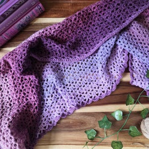May include: A handmade crochet shawl in shades of purple, transitioning from deep violet to lavender. The openwork design creates a delicate, lacy texture. The shawl is draped on a wooden surface, with a book and greenery in the background.