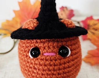 May include: A crocheted orange pumpkin with a black witch hat, large black eyes, and a small pink mouth. The pumpkin is a handmade item, perfect for Halloween or fall decor. The background includes fall leaves.