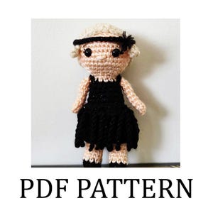 Puede incluir: Una muñeca de ganchillo con un vestido negro y una diadema con un lazo negro. La muñeca tiene la piel clara, mejillas rosadas y ojos negros. El texto "PDF PATTERN" se muestra debajo.
