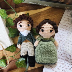 May include: Two handmade crochet dolls, a man and a woman, inspired by the novel "Pride and Prejudice." The man doll wears a green jacket, tan pants, and black boots. The woman doll is in a green dress. Both have brown hair and are on a wooden surface.