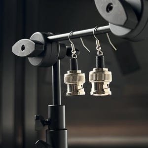 Może przedstawiać: Para srebrnych i czarnych kolczyków w kształcie złącz BNC, wiszących na czarnym stojaku.