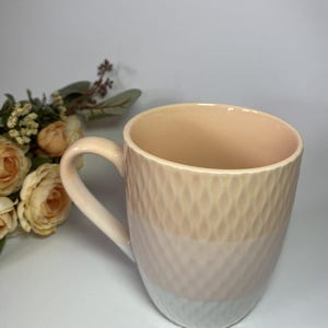 Peut inclure: Une tasse en céramique pêche avec un motif en losanges texturé sur la moitié supérieure. La partie inférieure est blanche et la poignée est de la même couleur pêche. La tasse est à côté d'un bouquet de roses crème.