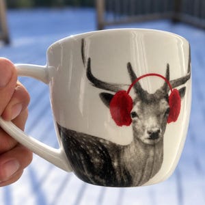 May include: A white ceramic mug featuring a black and white photographic print of a deer. The deer is depicted wearing bright red earmuffs with a thin red headband connecting them across the top of its head. The mug has a curved handle on the side.