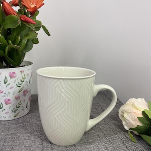 May include: A white ceramic mug with a black handle and the text "But First, Coffee" in a black, handwritten-style font. The mug is on a white surface.