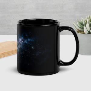 Peut inclure: Une tasse en céramique noire ornée d'un motif imprimé représentant une galaxie de l'espace profond avec des nébuleuses bleues et blanches tourbillonnantes et des étoiles dispersées sur un fond sombre. La tasse a une finition lisse et brillante.