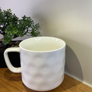 May include: A white ceramic mug with a textured, bumpy surface. The mug has a rounded shape and a handle.