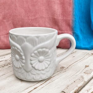 May include: A white ceramic mug featuring a raised, embossed owl design. The mug has a rounded body with detailed feather textures and large, circular eyes forming the central pattern. It includes a curved handle on the right side.