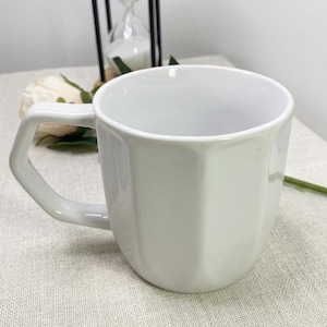 May include: A white ceramic mug with a geometric handle and a faceted body. The mug is empty and sits on a neutral-colored surface. An hourglass and a flower are in the background.