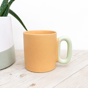 Peut inclure: Un mug cylindrique en céramique avec un corps de couleur pêche mate et une anse épaisse et arrondie de couleur vert menthe pâle. Le mug présente une finition lisse et unie, et est positionné debout, mettant en valeur son design simple.