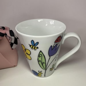 Peut inclure: Mug en céramique blanche décoré de fleurs, d'abeilles, d'une coccinelle et d'un escargot de dessin animé colorés. La tasse a une anse incurvée et une large ouverture. Le design présente des éléments jaunes, bleus, rouges, violets et verts.