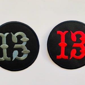 May include: Two circular patches with the number "13" embroidered on them. One patch features the number in a light blue and grey color, while the other has the number in red. Both patches have a black background.