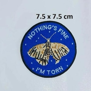 May include: A blue embroidered patch with a butterfly and the text "Nothing's Fine I'm Torn". The patch is 7.5 x 7.5 cm.