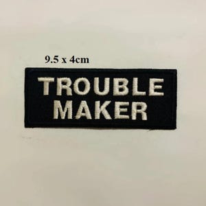May include: Black embroidered patch with white text that reads "Trouble Maker". The patch is 9.5 inches by 4 inches.