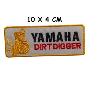 Pode incluir: Patch bordado, medindo 10 x 4 cm, com fundo branco e borda dourada. O patch apresenta o logotipo da Yamaha em preto e as palavras "DIRT DIGGER" em vermelho. Um gráfico de motocicleta dourada está à esquerda.