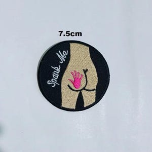 May include: A black and white embroidered patch with a nude figure and the text "Spank Me". The figure has a pink hand print on its backside. The patch is 7.5 cm in diameter.