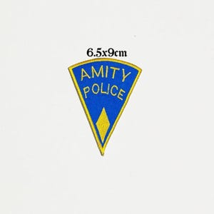 May include: A blue and yellow embroidered patch with the text "AMITY POLICE" in a triangular shape. The patch measures 6.5 x 9 cm.