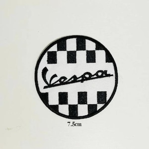 Puede incluir: Parche circular blanco y negro a cuadros con la palabra "Vespa" en una fuente de letra negra. El parche tiene un diámetro de 7,5 cm.