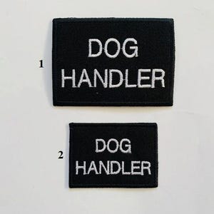 Può includere: Due toppe rettangolari nere con testo ricamato bianco. La toppa superiore, più grande, recita "DOG HANDLER". La toppa inferiore, più piccola, recita anch'essa "DOG HANDLER".