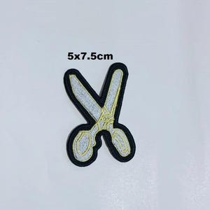 Puede incluir: Parche bordado de tijeras, que mide 5 x 7,5 cm. El parche presenta un diseño de tijeras plateadas con detalles dorados y un borde negro. El texto "5x7.5cm" está impreso encima de las tijeras.
