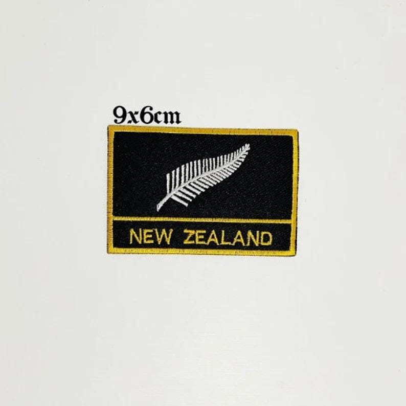 Op de afbeelding: Een rechthoekige zwarte patch met een gouden rand, met een witte varen en de tekst "NEW ZEALAND" in goud. Afmetingen: 9x6cm. Deze patch is waarschijnlijk een embleem of souvenir.