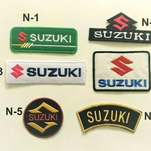 May include: Collection of six embroidered Suzuki patches in various shapes and colors. The patches feature the Suzuki logo and text in white, red, and yellow on green, black, and white backgrounds. The patches are rectangular, square, and circular.