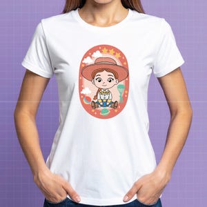 May include: White t-shirt featuring a cartoon illustration of a character wearing a cowboy hat. The character is inside an oval frame with stars, clouds, and planets. The design is in shades of red, orange, and blue.