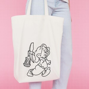 Puede incluir: Bolso tote de color crema con una ilustraci&oacute;n en blanco y negro de un personaje de dibujos animados enano. El enano corre llevando un pico y una linterna. El bolso tiene dos asas.