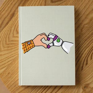 May include: A hardcover notebook with a light green cover. The cover features a cartoon illustration of two hands forming a heart shape. One hand is a human hand with a yellow plaid shirt sleeve, and the other is a robot hand.